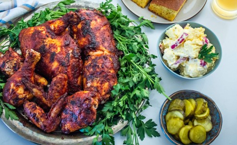 Recipe Blog - Classic BBQ Chicken - Top down sauced chicken on a large platter with green herbs, pickles, potato salad, cornbread, corn on cob, on a white countertop