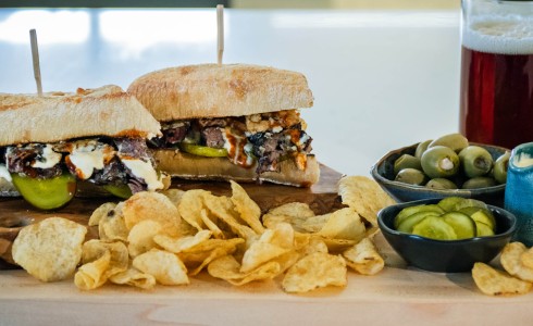 Recipe Blog - Father's Day Steak Sandwich - Large sandwich on light colored board with chips, olives, pickles, cheese sauce and brown ale on a white countertop