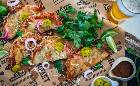 Recipe Blog - Pork Belly Tacos - Top down crispy tacos on a board with parchment topped with onion, jalapeno, and cheese, surrounded by limes and cilantro