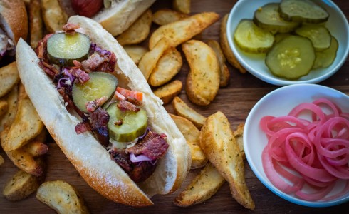 Recipe Blog - Great White North Dog - Top down hot dog topped with coleslaw, pickles, maple bacon on a board, surrounded by potato wedges, and pickles 