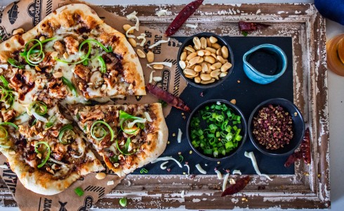 Recipe Blog - Kung Pao Pizza - Top down pizza on a rustic black and cream board on a white countertop with cheese, peppers, and navy napkins