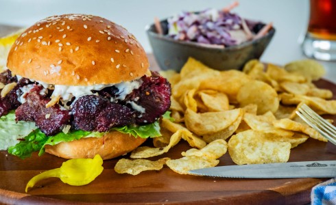 Recipe Blog - Beef Cheeks - Burger bun sandwich on a wood board with chips, a bowl of coleslaw, beer and cutlery, blue check napkin, white countertop