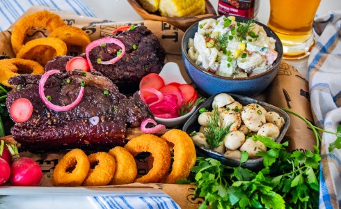 Recipe Blog - Dino Ribs - Top down ribs on a tray with pickled mushrooms, pasta salad, and onion rings with parsley and blue striped napkins and beer