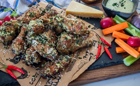 Recipe Blog - Garlic Parmesan Wings - Chicken wings on brown parchment on a slate tile on a wood board on a white countertop with blue and white check napkins, vegetables, and dip