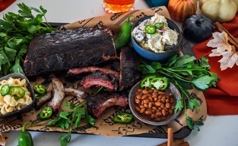 Recipe Blog - Pumpkin Spice Ribs - top down, White countertop, Orange napkins, rack of ribs on a tray surrounded by fresh parsley, macaroni, beans, and potato salad, pumpkins and cinnamon sticks adorn